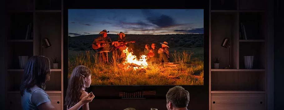 A family watches a movie of people around a campfire displayed on a large TV. The room is dimly lit, creating a cosy, intimate atmosphere.